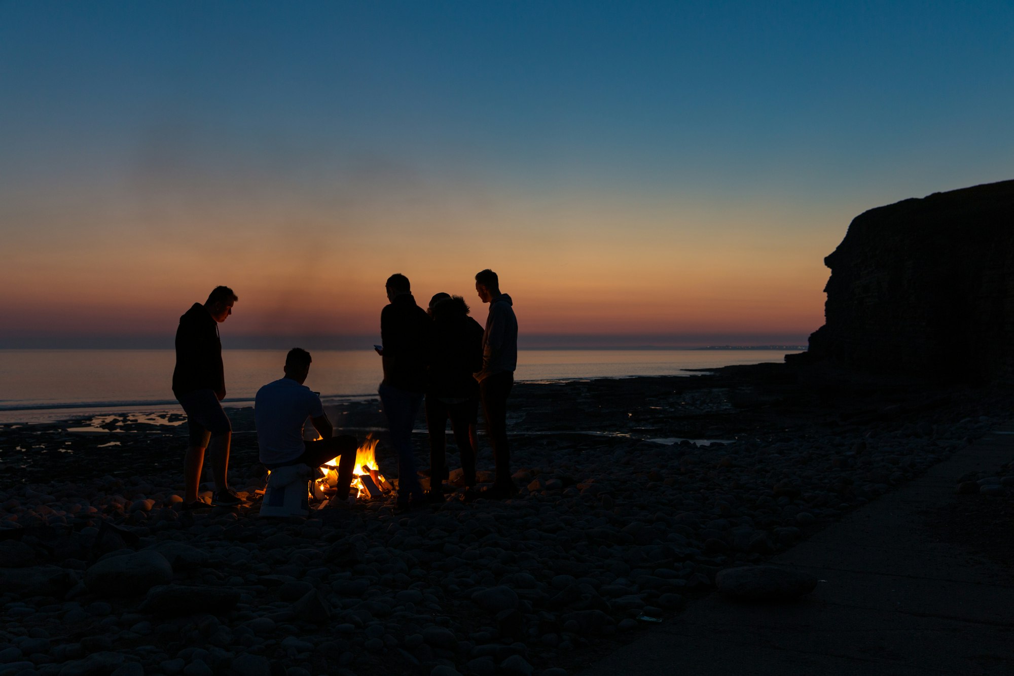 Mennesker samlet om et lille bål på en strand i skumringen.