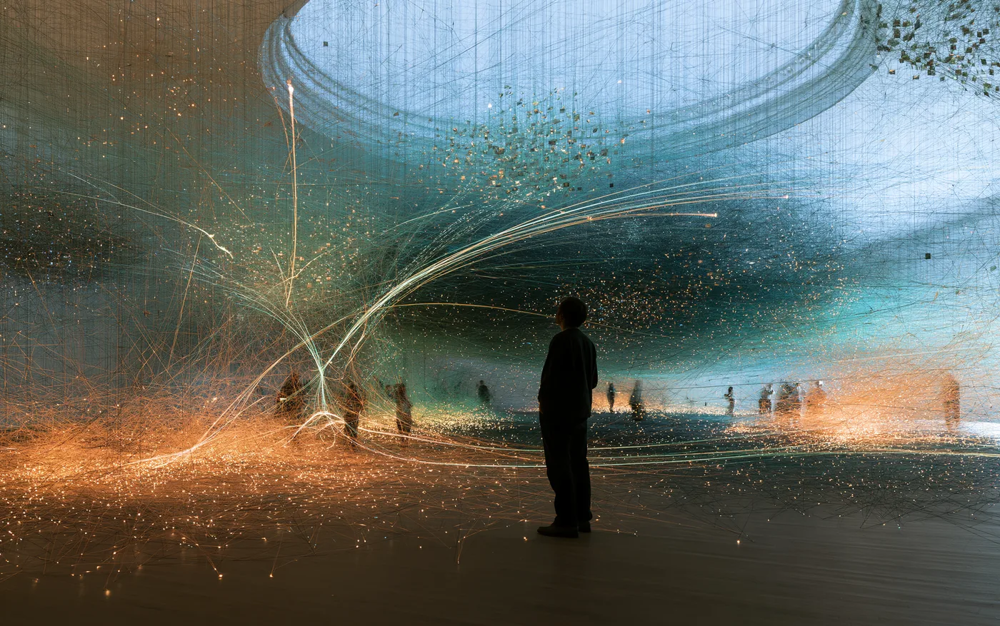 A person stands in a dark hall crossed by thousands of glowing threads and sparks, other people far off in the distance.